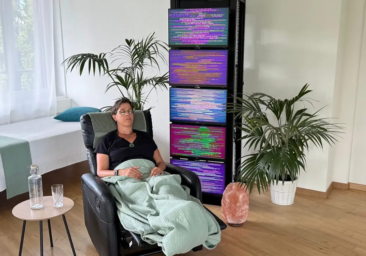 Eine Frau ruht mit geschlossenen Augen unter einer grünen Decke im Relaxsessel, daneben ein Turm mit fünf farbig leuchtenden Bildschirmen und Palmen.