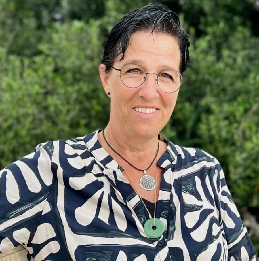 Porträt von Nicole Magrini-Mader mit Brille, kurzen Haaren und gemusterter Bluse vor grünen Büschen.