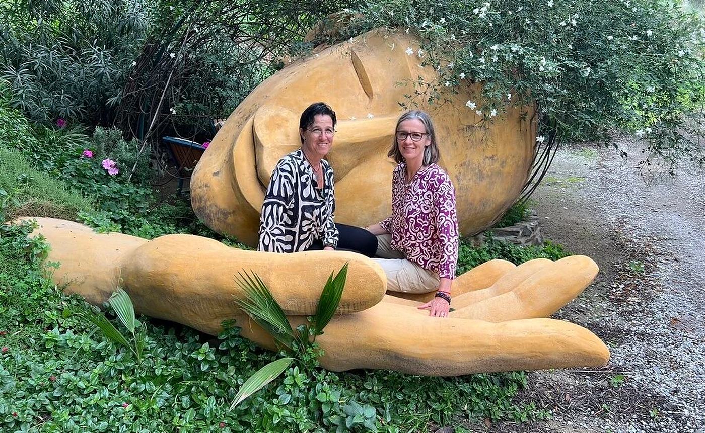 Sandra und Nicole Magrini-Mader sitzen lachend in einer grossen, aus Holz geschnitzten Hand im Grünen.
