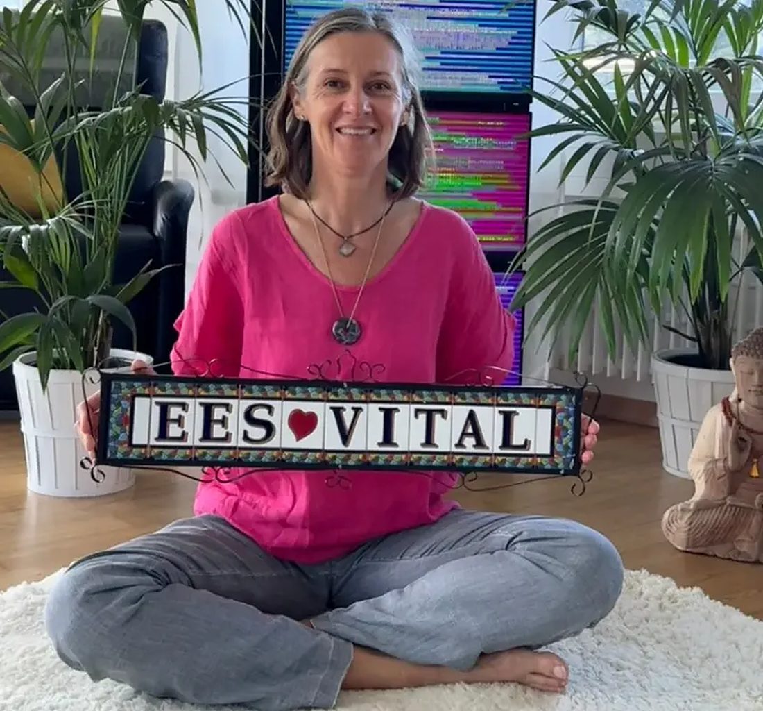Sandra sitzt im Schneidersitz vor einem Bildschirmturm und hält lächelnd ein Schild mit der Aufschrift EES Vital.