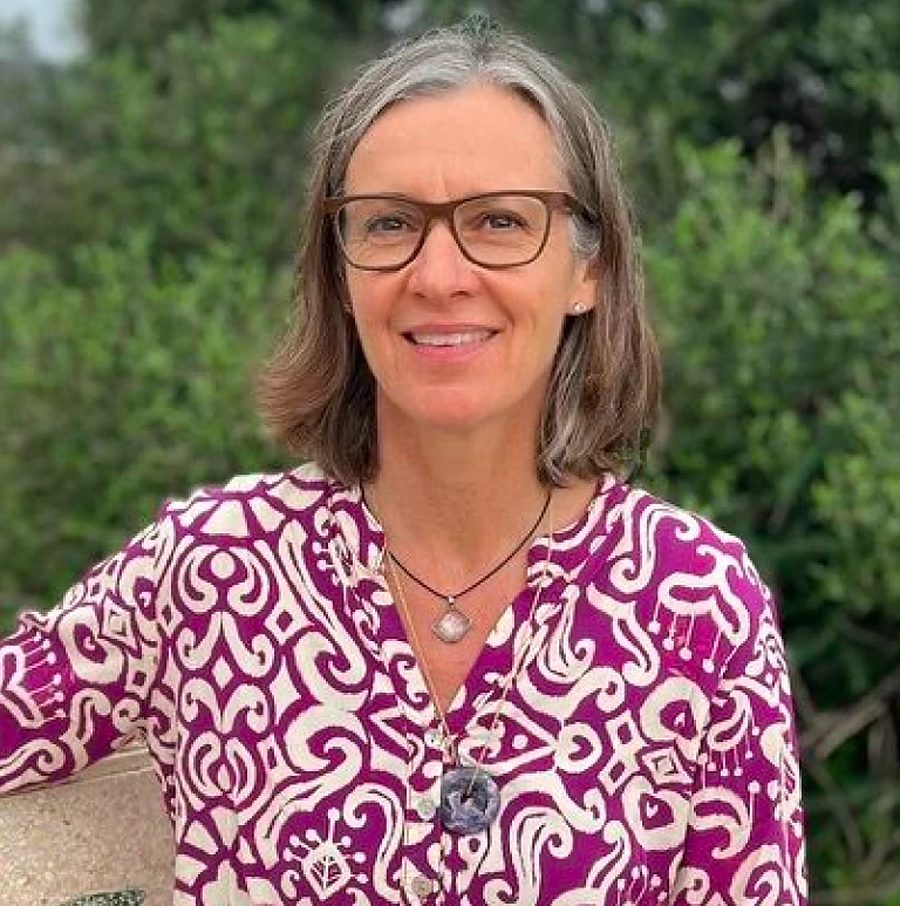 Porträt von Sandra Magrini-Mader mit Brille und gemustertem Oberteil vor grünen Büschen.